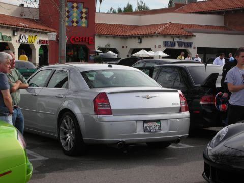1998 Ford Crown Victoria - silver Chrysler 300 sedan parked at a car show in front of storefronts including a Subway and a coffee shop.