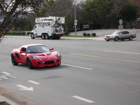 1998 Ford Crown Victoria - red Lotus Elise drives on a road, with a white utility bucket truck and a beige sedan in the background.
