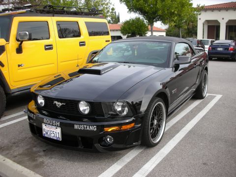1998 Ford Crown Victoria - black Roush Mustang convertible with hood scoops and a California plate sits next to a yellow Hummer H2 in a parking lot.