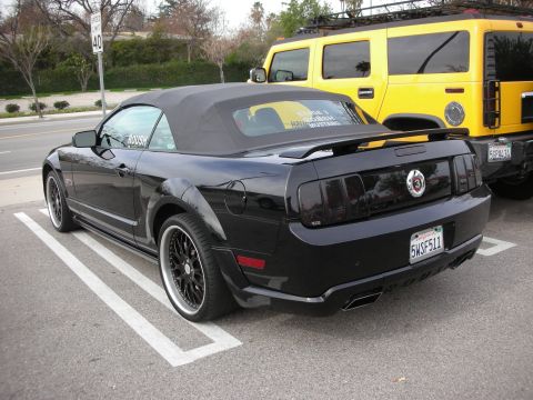 1998 Ford Crown Victoria - black Mustang convertible with rear spoiler and California plate parks beside a yellow Hummer H2 along a roadside lot.