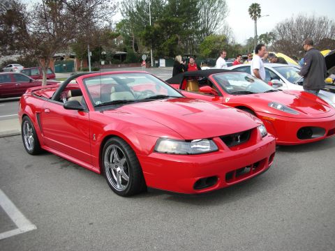 1998 Ford Crown Victoria - red Ford Mustang Cobra convertible with chrome wheels parks beside a red Ferrari coupe at a busy outdoor car gathering.