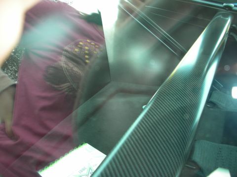 1998 Ford Crown Victoria - glare-filled close-up shot through glass shows a glossy carbon-fiber panel and a person in a maroon shirt reflected behind.