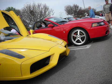 1998 Ford Crown Victoria - angular front of a yellow Ferrari Enzo sits beside a red Mustang convertible in a parking lot, spectators watching nearby.