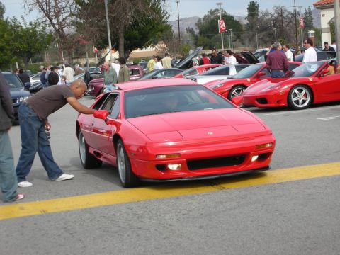 1998 Ford Crown Victoria - man wipes down a red Lotus Esprit in a parking lot at a car meet, with red Ferraris and a crowd in the background.