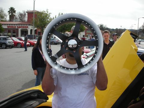 1998 Ford Crown Victoria - man in a white Cosworth shirt holds a black five-spoke alloy wheel up over his face beside a yellow car at a car show.