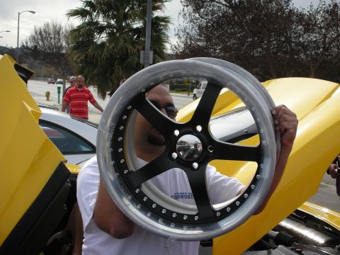 1998 Ford Crown Victoria - man holds a black five-spoke alloy wheel with a polished lip up to his face next to an open-door yellow car at a meet.