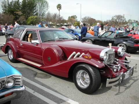 1998 Ford Crown Victoria - maroon neoclassic car with chrome external exhaust pipes, side-mounted spare, and wire wheels parked at a car show.