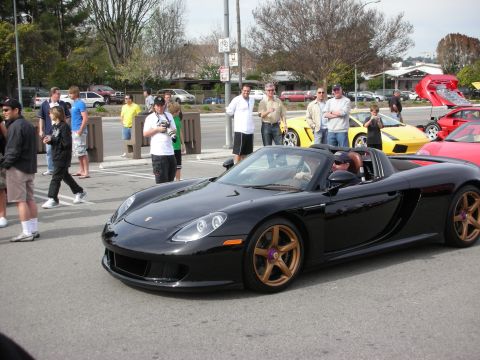 1998 Ford Crown Victoria - black Porsche Carrera GT with gold wheels is driven slowly through a car meet as a crowd looks on.