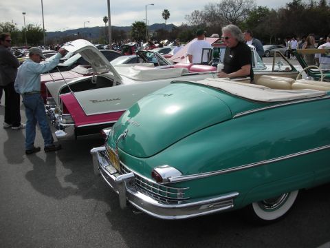 1998 Ford Crown Victoria - teal Packard convertible parked beside a pink-and-white one in a row of classic cars at an outdoor car show.