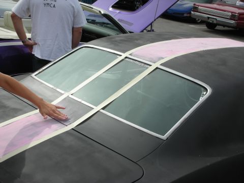 1998 Ford Crown Victoria - person's hand rests on a matte-black roof being prepped for twin stripes, with pink primer and masking tape laid out at a car show.
