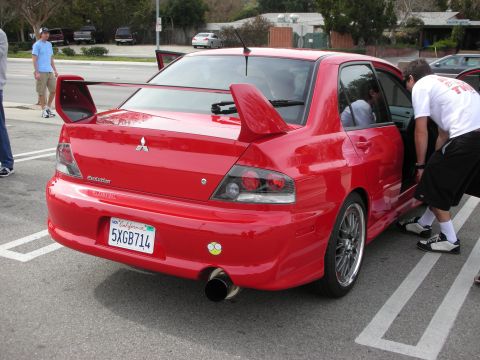 1998 Ford Crown Victoria - Rear three-quarter view of a red Mitsubishi Lancer Evolution sedan with a tall rear wing as people stand nearby in a parking lot.