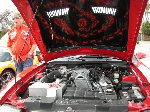 1998 Ford Crown Victoria - man in a red Ferrari shirt stands by a red car with its hood up, revealing flame airbrush art on the underside and a V8 engine.