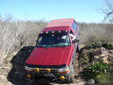 1995 Ford Bronco - This red 1995 Ford Bronco, equipped with a red camper shell, navigates an off-road trail under a clear blue sky.