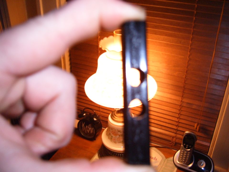 Black plastic part with two circular holes, held by a finger. A lamp and a cordless phone are visible in the background