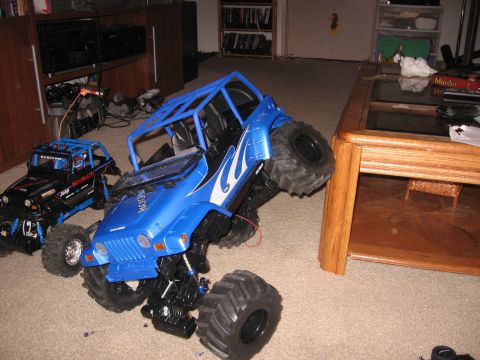 1990 Ford Bronco - A blue remote control monster truck is positioned on a carpeted floor next to a black RC vehicle.