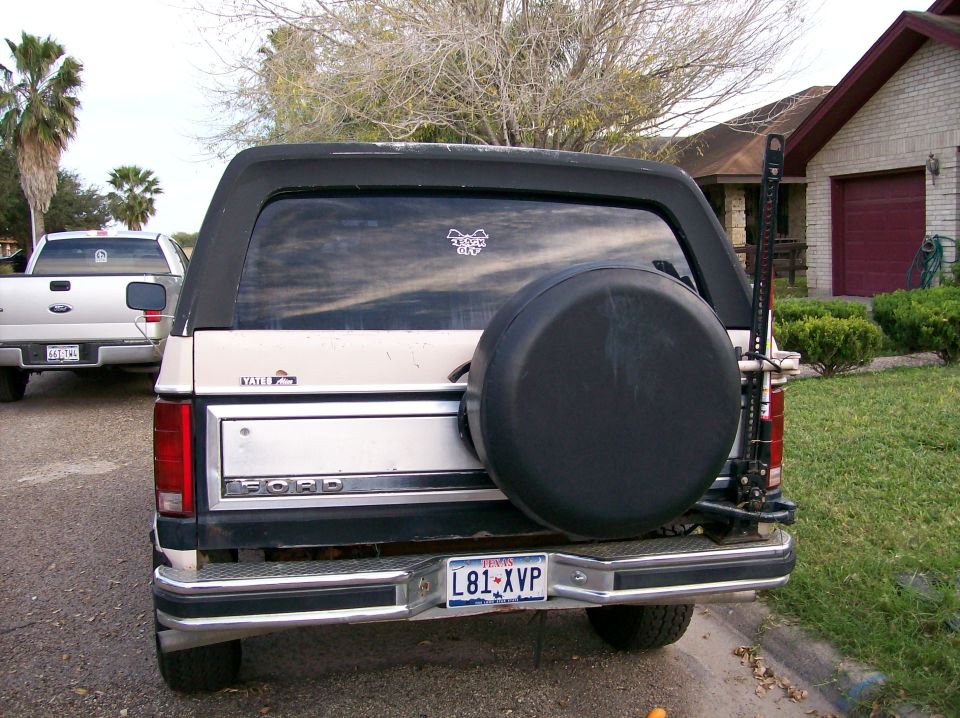 Tan Ford Bronco with black top and spare tire on rear, parked next to a white pickup truck