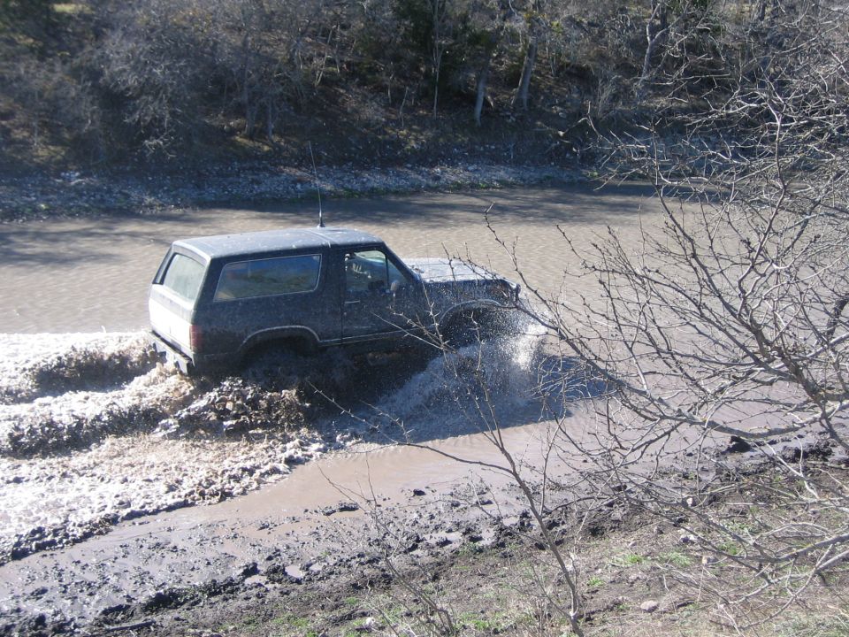 Dark colored Ford Bronco driving through a muddy stream, kicking up water
