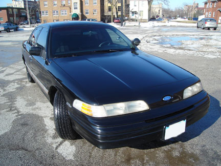 1992 Ford Crown Victoria - A black 1992 Ford Crown Victoria sedan is parked outdoors on a sunny day, with buildings in the background.