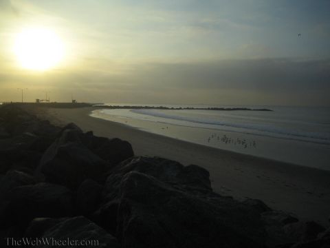1990 Ford Bronco - Coastal beach scene at dawn with the sun low over the ocean, dark rocks in the foreground and a distant jetty.