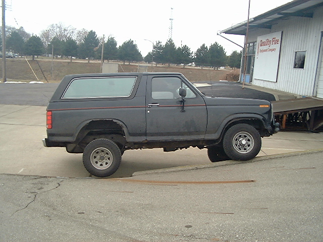 Black lifted two-door Ford Bronco in side view, muddy tires, parked by a white building.