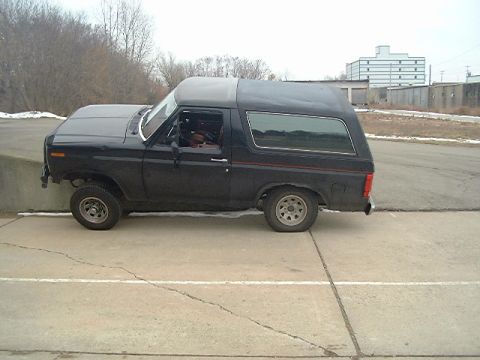 1986 Ford Bronco - A black SUV is parked on a concrete surface with a road and buildings in the background.