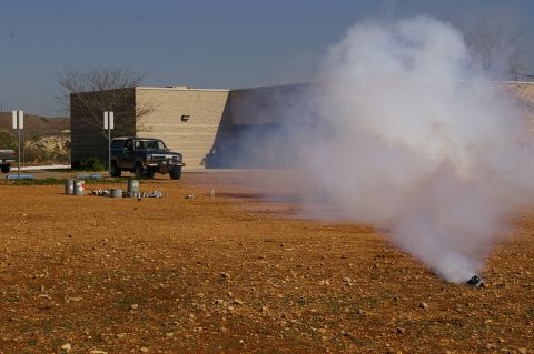 1986 Ford Bronco - A dark-colored SUV is parked in a lot with several cans scattered nearby, as a large plume of smoke obscures the scene.