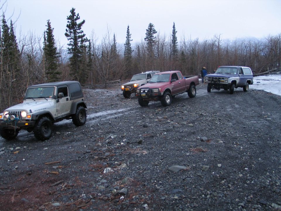 Jeep, pickup truck, and two SUVs on a gravel road with trees and mountains in background