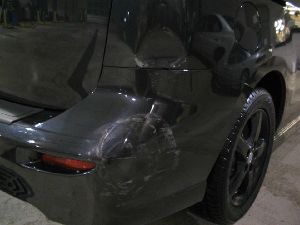 Black minivan rear bumper with significant damage, dented and scraped. Visible taillight and wheel