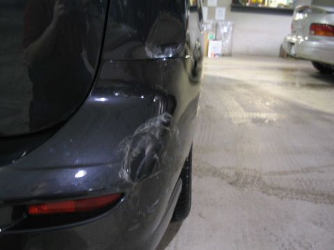 2006 Mazda Mazda 5 - Close-up of a damaged rear bumper on a dark vehicle, with another car partially visible in the background.