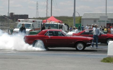 1963 Ford Fairlane - A red 1967 Ford Mustang performs a burnout at the drag strip, with other vehicles and spectators in the background.