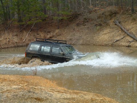 1994 Ford Bronco - A black Jeep Cherokee XJ navigates through a shallow body of water, creating a large spray.