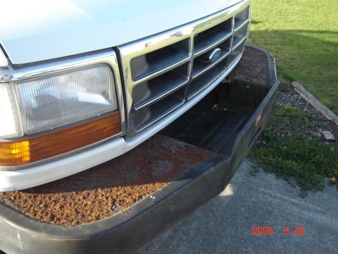 1993 Ford Bronco - The front grille of a white Ford Bronco shows a chrome emblem and horizontal bars, with a heavily rusted bumper below.
