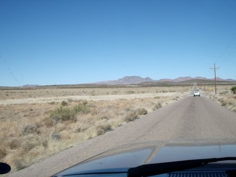 1996 Ford Bronco - Windshield view over a dark hood on a two-lane desert highway, with a white vehicle ahead, power poles along the shoulder and low mountains…