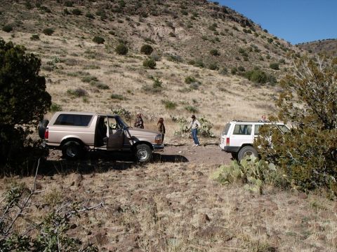 1996 Ford Bronco - Two SUVs are stopped on a dirt road in a dry, hilly landscape. The Ford Bronco's door is open, and people stand near the vehicles.