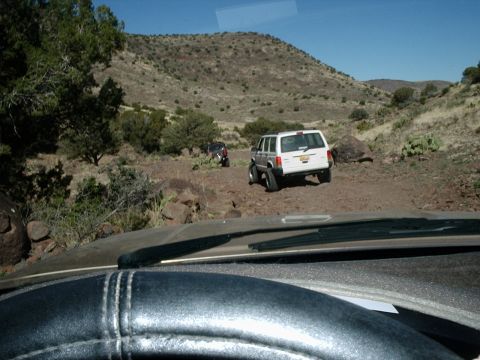 1996 Ford Bronco - Two SUVs navigate a rough dirt trail through a dry, hilly terrain under a clear blue sky.