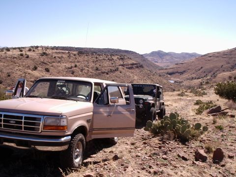 1996 Ford Bronco - Two off-road vehicles are parked on a dusty trail in a mountainous desert area under a clear blue sky.