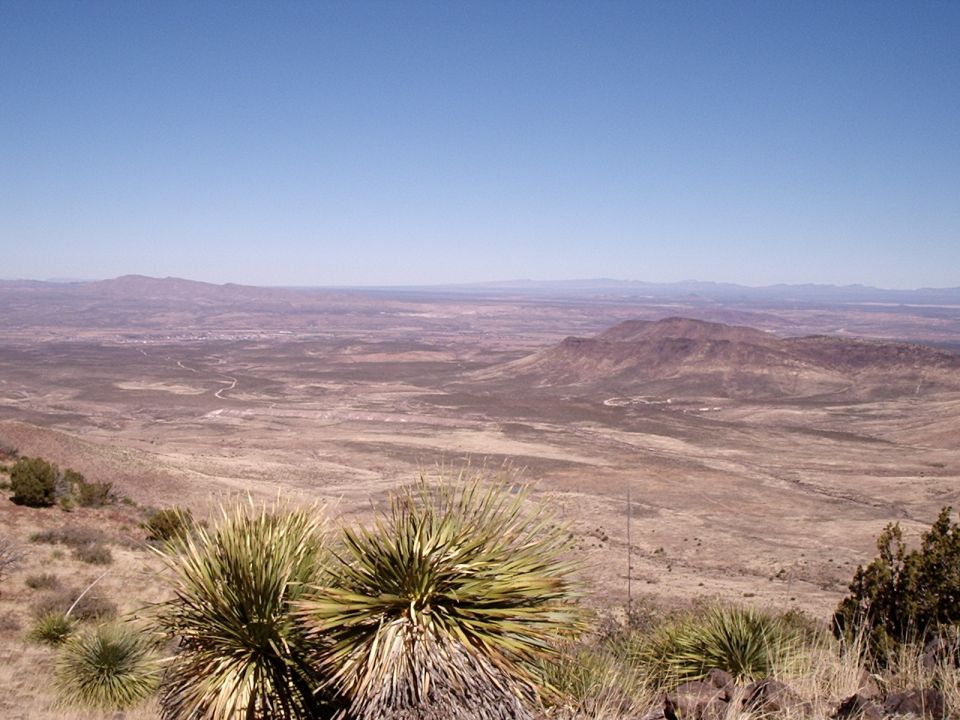 Desert landscape with rolling hills, distant mountains, and a small town visible in the valley under a clear blue sky