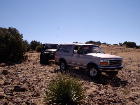 1996 Ford Bronco - Two off-road vehicles, a tan Ford Bronco and a black Jeep Wrangler, are parked on a dry, rocky trail under a clear blue sky.