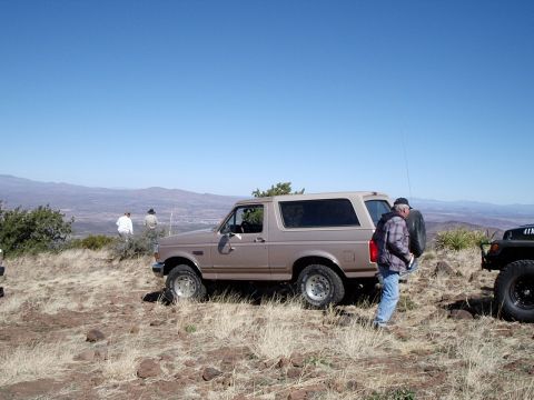 1996 Ford Bronco - A tan 1996 Ford Bronco is parked on a dry, grassy hillside overlooking a valley, with another vehicle partially visible to the right.