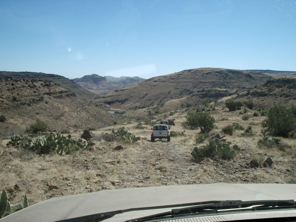 Two SUVs driving on a rocky dirt trail through a dry, hilly desert with cacti and sparse brush under a clear blue sky