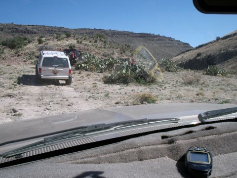 1996 Ford Bronco - Two off-road vehicles are parked on a dusty trail surrounded by desert vegetation and hills.