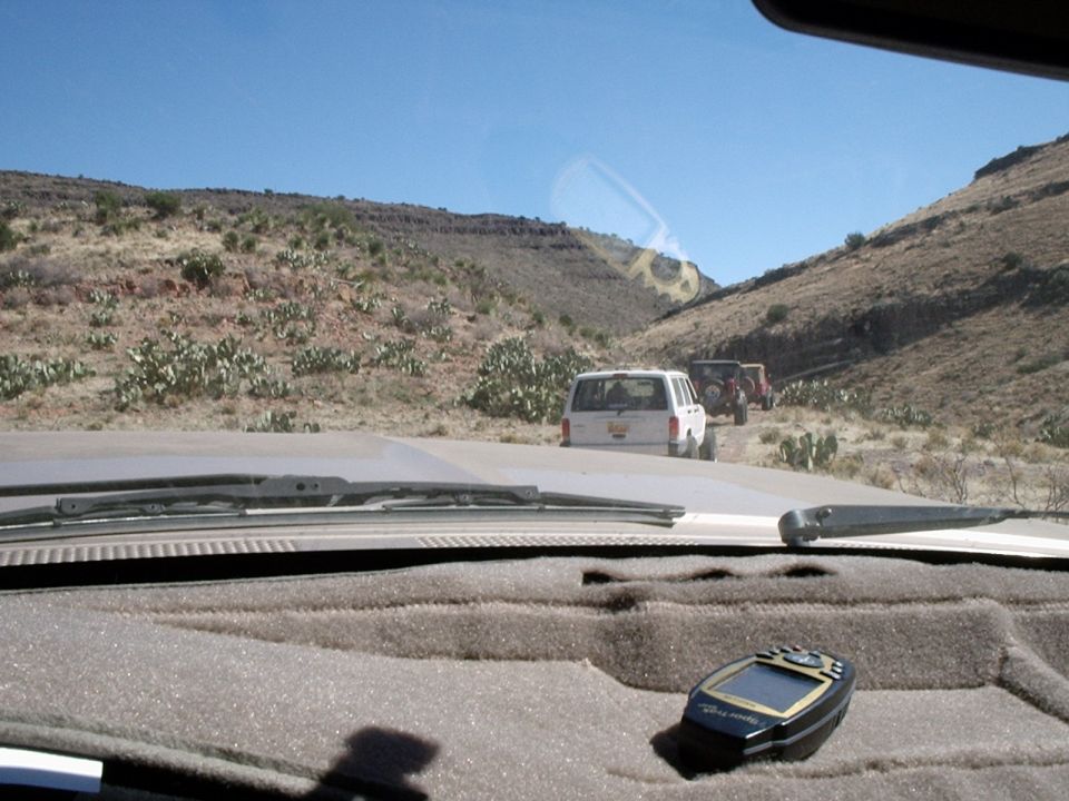 View from inside a vehicle looking at a line of SUVs on a dirt road in a desert with hills and cacti