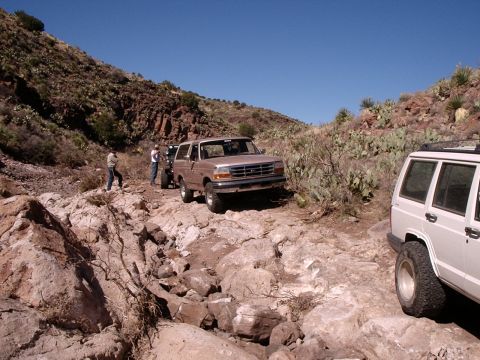 1996 Ford Bronco - Two SUVs, a 1996 Ford Bronco and a Jeep Cherokee, traverse a challenging rocky path in a desert landscape.