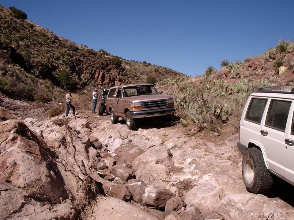 1996 Ford Bronco and white Jeep Cherokee navigating a rough, rocky off-road trail with sparse desert vegetation