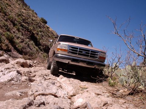 1996 Ford Bronco - A tan Ford Bronco navigates a challenging rocky terrain under a clear blue sky.