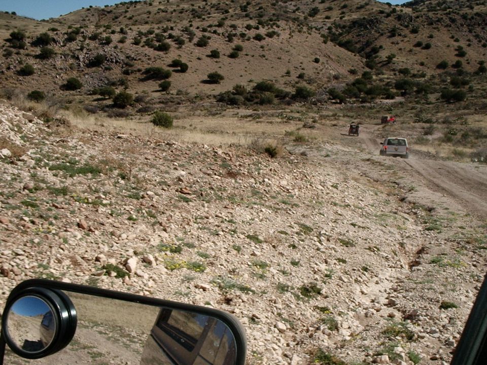 Several off-road vehicles driving on a dusty dirt road through a dry, hilly landscape. A side mirror is visible in the foreground