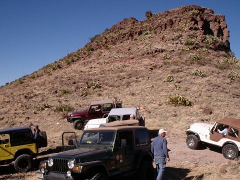 1996 Ford Bronco - A group of off-road vehicles are gathered on a dusty trail beneath a large, arid hill.