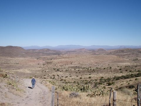1996 Ford Bronco - A lone figure walks down a dusty trail through a vast, arid landscape under a clear blue sky.