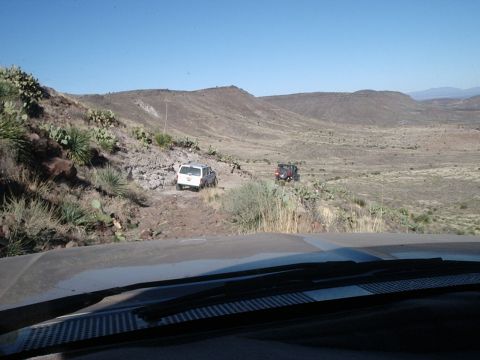 1996 Ford Bronco - Two off-road vehicles navigate a trail through a dry, hilly terrain under a clear blue sky.