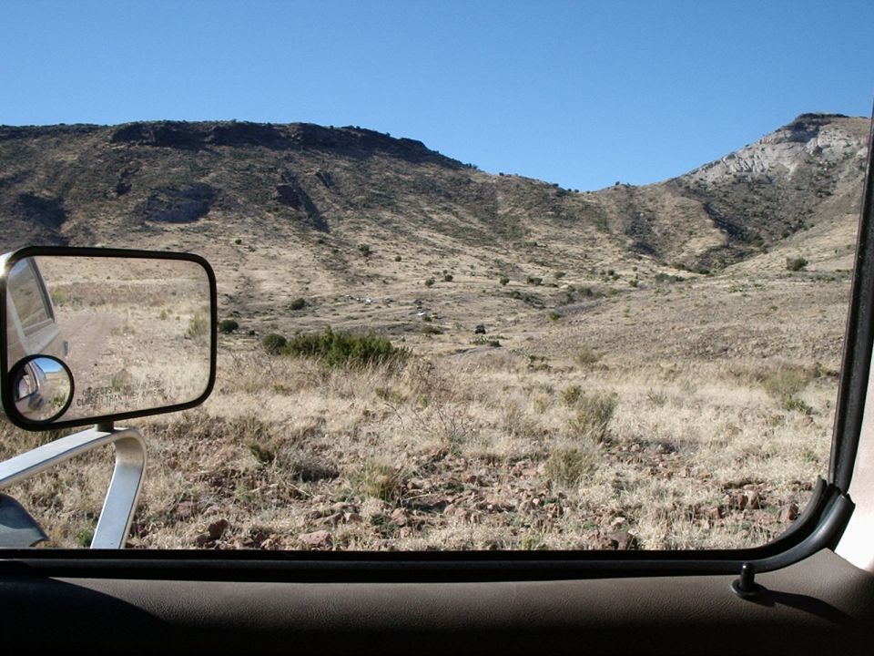 Desert hills and dry grass seen through a car window, with a side mirror reflecting the road